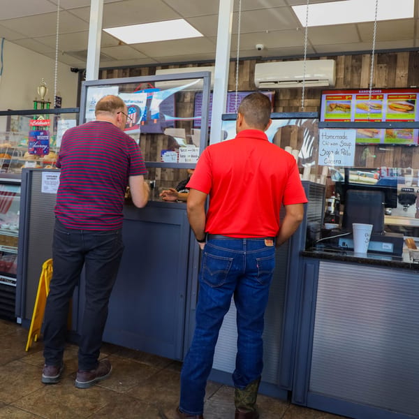 Two men ordering food at the counter of Tom's Cuban on the Go, with menu boards visible.