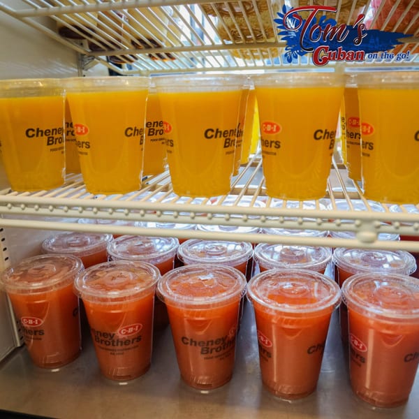 A refrigerated display case holds rows of orange and red juices in clear plastic cups.