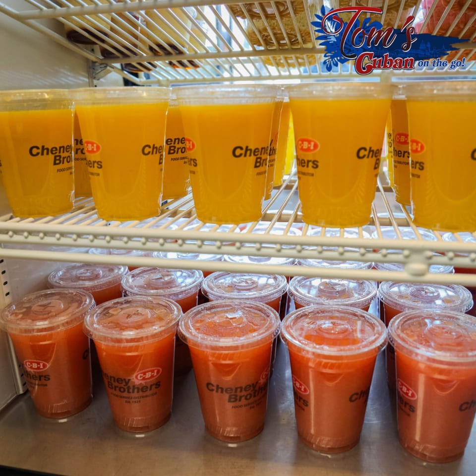 A refrigerated display case holds rows of orange and red juices in clear plastic cups.