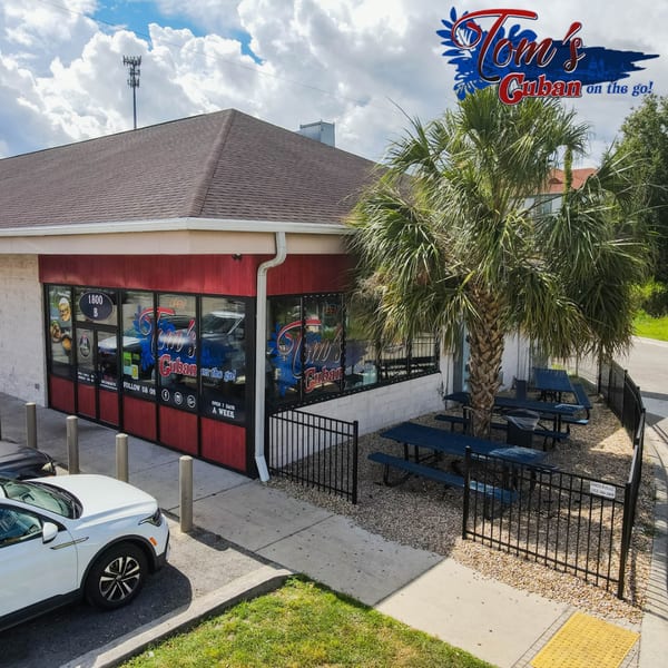 Exterior of Tom's Cuban on the Go restaurant with red accents, outdoor seating, and a palm tree.
