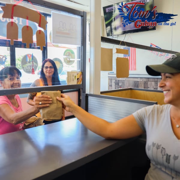 A smiling Tom's Cuban on the Go employee hands a customer a brown paper bag through a window.