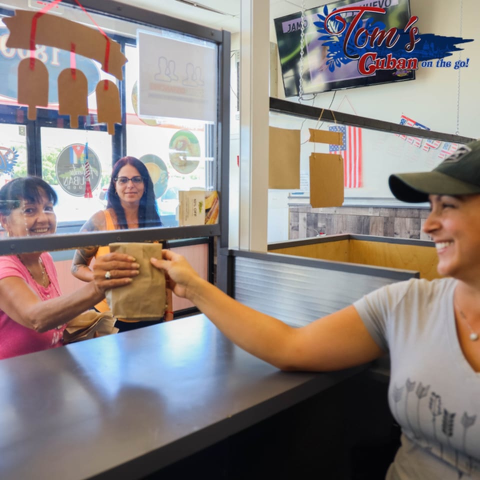 A smiling Tom's Cuban on the Go employee hands a customer a brown paper bag through a window.