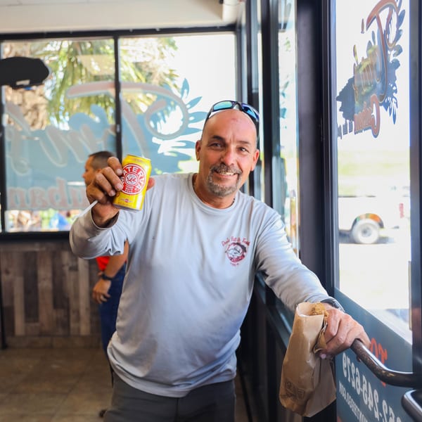 A man in a light blue shirt smiles, holding up a yellow Yerba Mate soda can and a brown paper bag.