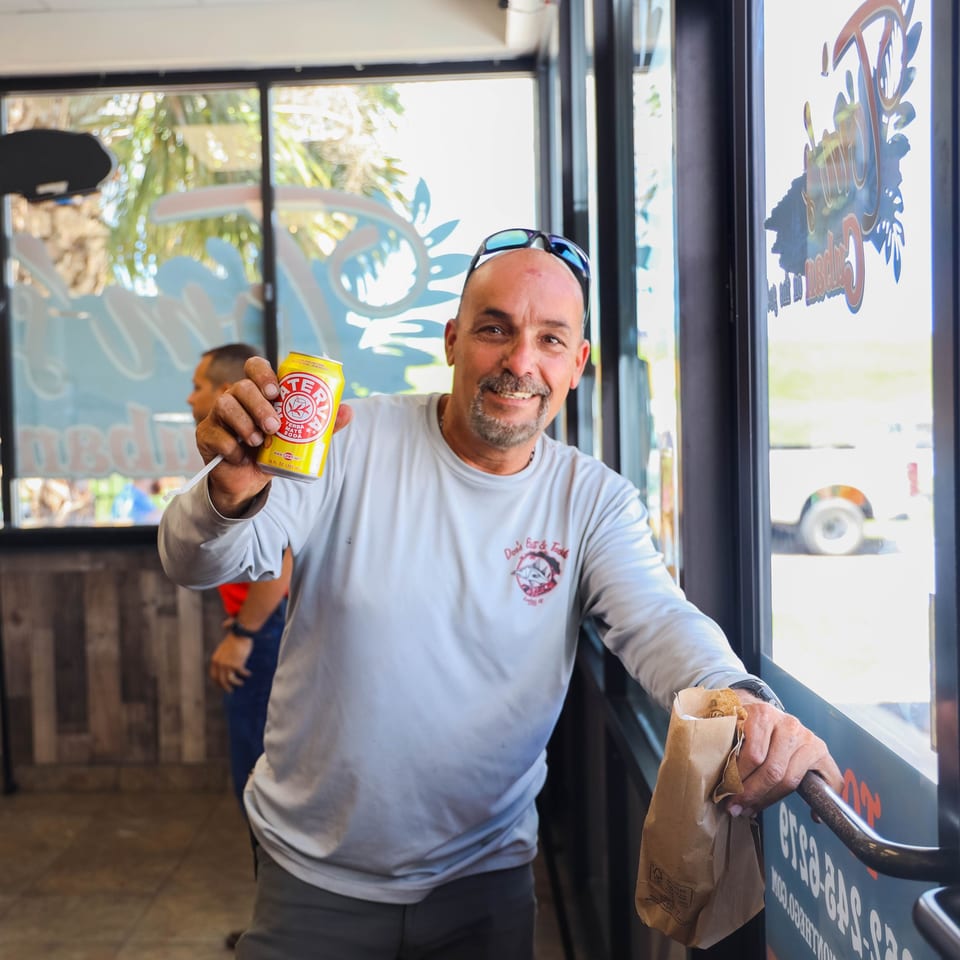 A man in a light blue shirt smiles, holding up a yellow Yerba Mate soda can and a brown paper bag.