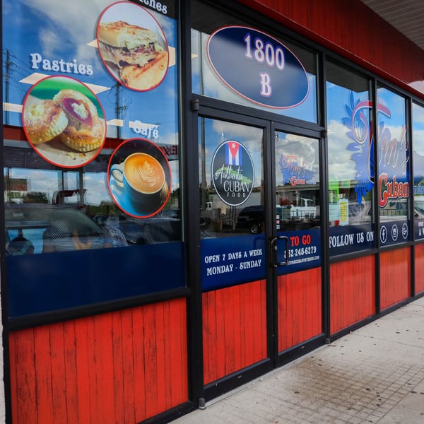 Exterior of Tom's Cuban on the Go, with window decals showing Cuban sandwiches, pastries, and coffee.