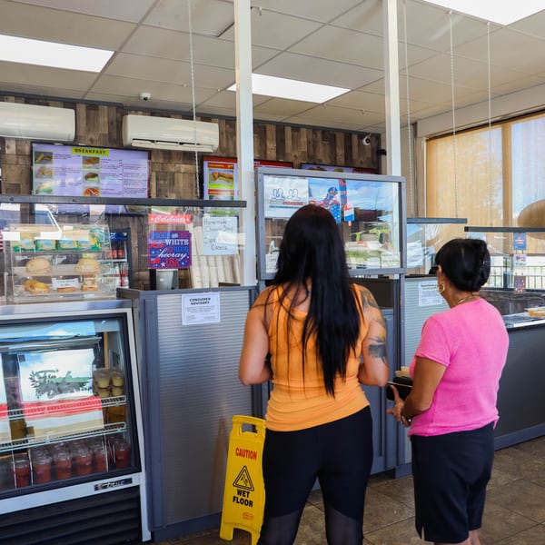 Two women ordering Cuban food at a counter with a breakfast menu and pastries on display.
